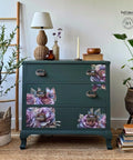 A vibrant, nature-themed room features a dark green dresser adorned with detailed floral and butterfly decals. The dresser is flanked by plants in woven baskets and accented with a lamp, vases, and decorative items on top. A wooden ladder stands to the left.