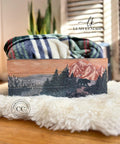 A cozy and artistic scene featuring a beautifully hand-painted wooden box with a mountainous landscape, positioned on a soft, fluffy white surface with a plaid blanket gently draped over it, evoking a sense of warmth and homeliness using the National Parks A3 Rice Decoupage Paper.