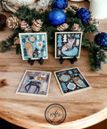 Holiday-themed coasters featuring winter scenes with animals are displayed on a wooden table. Two coasters are standing on small black easels, and two are laid flat. Pine and blue ornament decorations are in the background. A circular logo is present at the bottom.