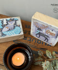 Two small, square wooden blocks with winter-themed illustrations sit on a rustic wooden table. One block features a fox in a snowy forest, and the other shows a reindeer and a house. A lit candle in a dark holder and a sprig of greenery are in the foreground.