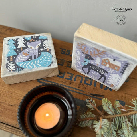 Two small, square wooden blocks with winter-themed illustrations sit on a rustic wooden table. One block features a fox in a snowy forest, and the other shows a reindeer and a house. A lit candle in a dark holder and a sprig of greenery are in the foreground.