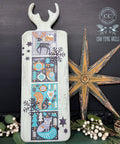 A rectangular board with a handle and antler-shaped top displays a vibrant winter-themed design featuring a stag, owl, squirrel, and fox among trees and snowflakes in shades of blue, green, and orange. Nearby is a large rustic star and clusters of white berries.