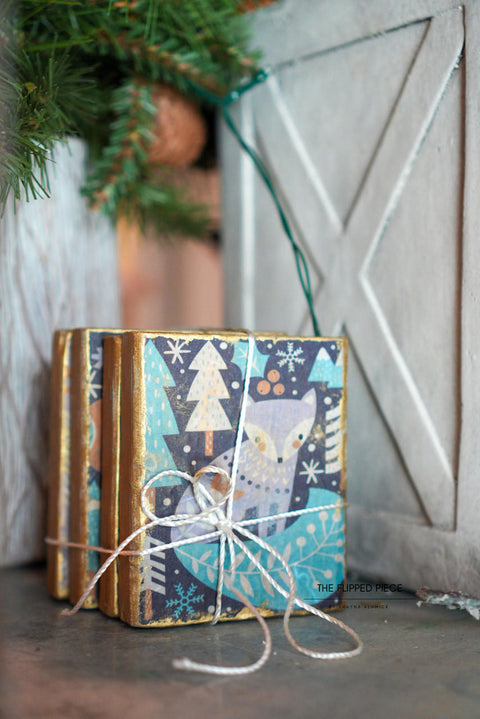 Gold blocks wrapped adorned with the Nordic Noel Decoupage Paper with snowflakes and trees, tied with a white string. Background features subtle, rustic decor including a wooden surface and green pine branches.