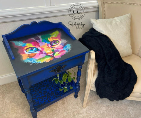A small, ornate blue table with a colorful, abstract painting of a cat's face on its top. The bottom shelf has a blue leopard print design and holds a small plant. The table is next to a beige armchair with a white pillow and a fluffy black blanket draped over it.
