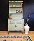 A vintage green cabinet with ornate details stands against a dark wall. It displays a decorative box, books, and shiny cups. Below, a large white vase with twisted branches sits to the right. A rug with floral patterns lies on the wooden floor.