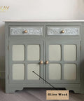 A light green cabinet with intricate patterns on the top drawers and gold knobs. "Olive Wood" is labeled with an arrow pointing to a section. The cabinet stands on a wooden floor with a potted plant nearby.
