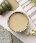 A jar filled with green paint, a green massage tool, a greenish fabric patch, a striped cloth, and white flowers are artistically arranged on a white surface. The scene has a natural and calming aesthetic.