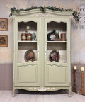 A vintage green cabinet with glass-paneled doors displays decorative plates, books, and ornaments. It’s topped with greenery. The wall features framed art and a watermark for “Low Flying Angels” with a winged design and “Dixie Belle Paint Company Content Creator” . Candlesticks sit nearby.
