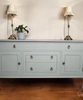 A light blue vintage sideboard with brass handles stands against a cream wall, flanked by two ornate table lamps with white shades. A small potted plant sits on top, and the floor is wooden.