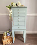 A tall, light blue wooden chest with seven drawers stands against a beige wall, decorated with white and pale flowers on top. Wicker baskets with more flowers are arranged on the floor beside it.