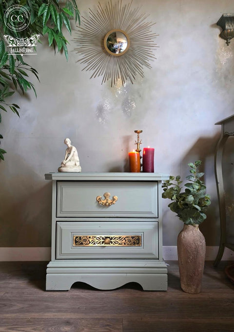 A small sage green dresser with two drawers sits against a gray wall. On top are a white statue, two lit candles, and a gold candlestick. A round sunburst mirror and leafy plant decor are in the background. A large vase with leaves is beside the dresser.