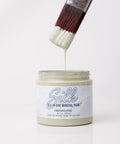A hand holds a paintbrush dripping light grey paint over an open jar labeled "Silk All-In-One Mineral Paint Oxford Fog" against a plain white background.
