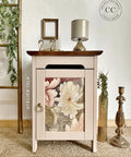 A small wooden cabinet with a floral design on the door sits on a woven rug. Atop the cabinet are two swan figurines and a small gold lamp. A golden frame hangs on the wall above. A wooden ladder with a plant and a gold candle holder are nearby.