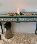 A blue-green vintage console table with intricate white filigree designs, gold knobs, and two drawers. A lit candle, beaded garland, and greenery decorate the top. The table sits on a fluffy rug, next to a glass jar with a plant.