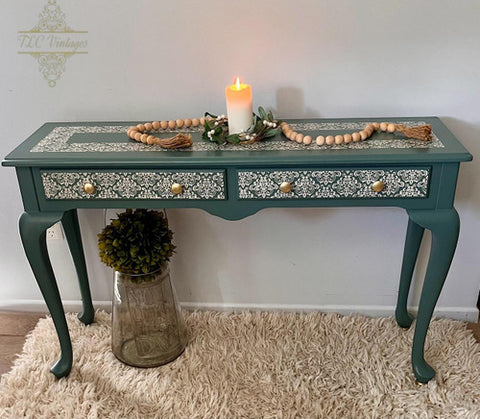 A blue-green vintage console table with intricate white filigree designs, gold knobs, and two drawers. A lit candle, beaded garland, and greenery decorate the top. The table sits on a fluffy rug, next to a glass jar with a plant.