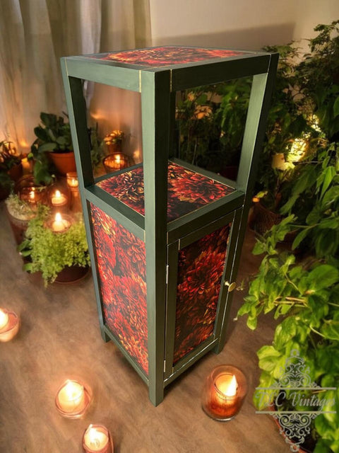 A small green wooden cabinet with red floral patterns on the door, shelf, and top, surrounded by lit candles and potted plants in a cozy, softly lit room.