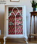 White cabinet with floral pink and red decoupage paper, displaying various items inside. It stands next to a wooden plant stand holding a green plant. The scene is set against a wooden floor and light-colored wall with a watermark.