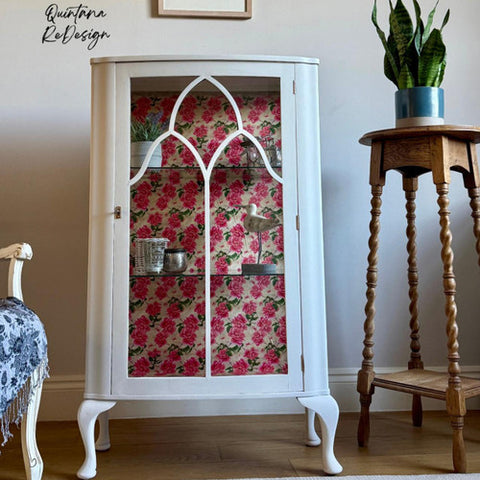 White cabinet with floral pink and red decoupage paper, displaying various items inside. It stands next to a wooden plant stand holding a green plant. The scene is set against a wooden floor and light-colored wall with a watermark.