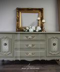 A vintage light green dresser with ornate gold handles sits against a wall. On top are a gold picture frame, a bouquet of white flowers, and two gold candlesticks. Gold curtains hang beside a window with blinds.