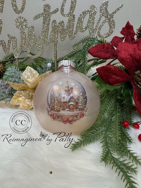 A Christmas ornament with a snowy village scene is displayed among pine branches, red poinsettias, and a golden "Christmas" sign in the background. The ornament features a church and trees dusted with snow.