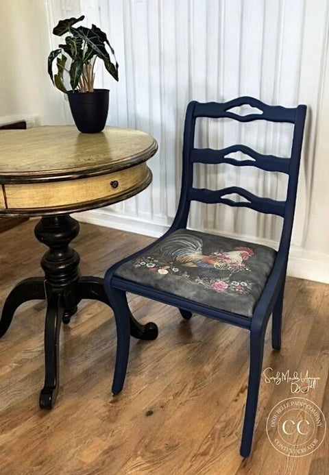 A vintage wooden chair painted in navy blue with a cushion featuring a colorful rooster and floral design sits beside a round, antique dark wooden table. A potted plant with variegated leaves rests on the table, set against a white paneled wall.