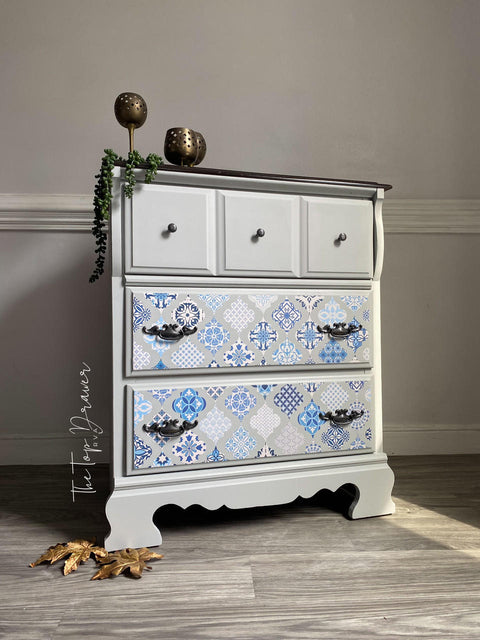 A gray 3 drawer dresser. Sapphire Sonata Transfer is seen applied to the bottom two drawers, the bottom two drawers have long intricate hardware, the top drawer has knobs hardware. Staged with bronze cups with a vine  on top of the piece and large gold leaves on the floor.