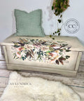 A wooden chest, decorated with an intricate floral design featuring roses and foliage, is placed against a white wall. A green textured pillow rests on top of the chest. A fluffy rug lies in front.