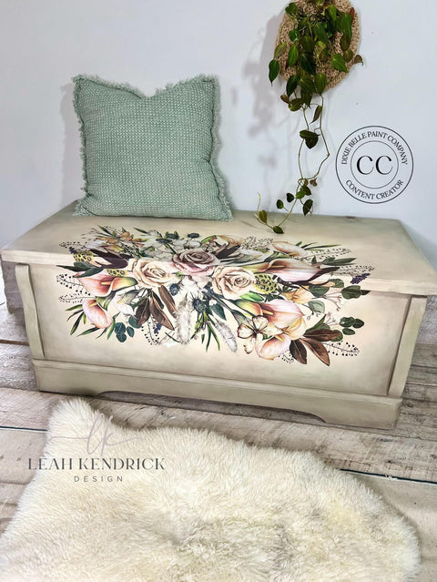 A wooden chest, decorated with an intricate floral design featuring roses and foliage, is placed against a white wall. A green textured pillow rests on top of the chest. A fluffy rug lies in front.