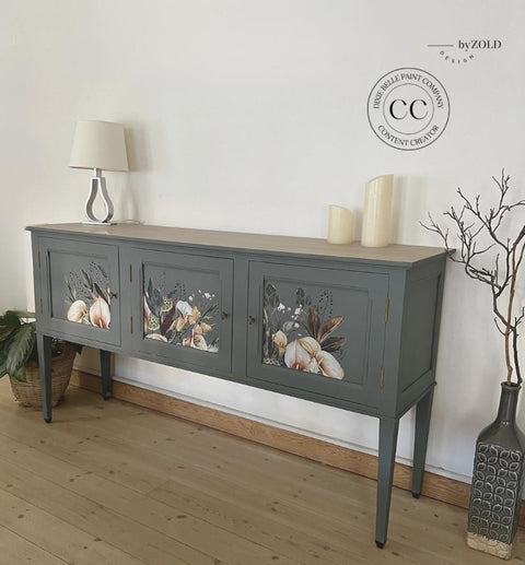 A vintage-style sideboard painted in dark grey with floral designs on its doors. It is positioned against a plain white wall. The sideboard is decorated with a lamp and two candles.