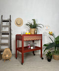 Red painted drink cart with black wheels. Staged with glasses, lemons, a shaker cup, a candle inside a vase and a wicker basket containing faux greenery on top.