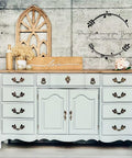 Baja Gray Silk All-In-One paint is perfect for painting kitchen cabinets, bathroom cabinets and painting furniture. Baja Gray Silk All-In-One Mineral Paint on a cabinet with bronze hardware. The piece is staged with a sign that says "Love" and a flower arrangement. The cabinets sits in front of a white brick wall.