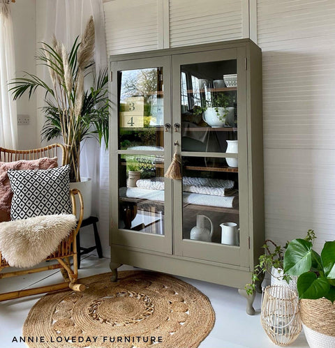 Hampton Olive Silk All-In-One paint is perfect for painting kitchen cabinets, bathroom cabinets and painting furniture.
Glass display case painted in Hampton Olive Silk All-In-One paint staged with boho decor around the piece like a brown woven rug, potted houseplants, and a chair with pillows and a fur blanket.