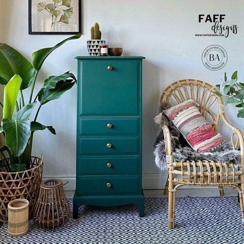 Midnight Green Silk All-In-One paint is perfect for painting kitchen cabinets, bathroom cabinets and painting furniture. Dark green Silk all In one painted tall dresser. Staged with some wicker potted plants a nd a boho style chair with pillows and blankets next to it. On top has a potted cactus, a wood bowl and candle.
