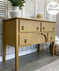 Mojave Silk All-In-One paint that is perfect for painting kitchen cabinets, bathroom cabinets and painting furniture. Dark Yellow desk with nailhead trim stencil for accent, and gold hardware. Staged with potted plant and a wicker basket.