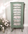 A vintage-style cabinet painted in green with six drawers. Each drawer features a floral and dragonfly pattern design in subtle colors. Two silver handles are on the side. The background includes white flowers on the left and a white wall.