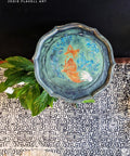 A round, intricately designed wooden tray features a painted image of a fox surrounded by blue floral patterns. The tray sits on a tiled surface with a plant nearby. Text in the top left corner reads "Jodie Flavell Art.
