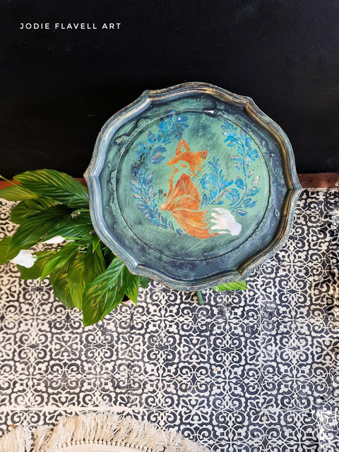 A round, intricately designed wooden tray features a painted image of a fox surrounded by blue floral patterns. The tray sits on a tiled surface with a plant nearby. Text in the top left corner reads "Jodie Flavell Art.