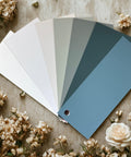 A fan of six paint color swatches ranging from off-white to blue-green is displayed on a beige surface, surrounded by small white flowers.