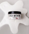 A round container labeled "Stick With Me Dixie Shine Glue 3.38 oz | 100 mL" sits on a white, glossy, spilled substance against a light background.