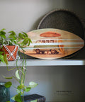 A decorative shelf displaying the Surf's Up Decoupage Paper on a small surfboard, accompanied by woven baskets, a potted plant in macrame, and a stack of books. The setting conveys a cozy, artistic vibe.