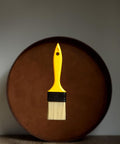 A yellow paintbrush with light bristles is suspended in front of a large, round, brown object against a plain background.