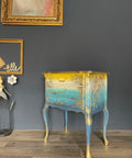Blue Moon Terra Clay Paint provides endless possibilities for your imagination and can be used on wood, canvas, tile, fabric and more!
Bedside table with ombre design going from yellow to a tan fading into a Cerulean Blue Terra Clay Paint blending in to  Blue Moon Terra Clay Paint. Using gold hardware and gold on woodwork. Staged In front of black wall and gold picture frames.