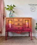 Bougainvillea Terra Clay Paint provides endless possibilities for your imagination and can be used on wood, canvas, tile, fabric and more!
Wet distressed credenza with Bougainvillea Terra Clay Paint on the bottom portion of the ombre with Daffodil Terra Clay Paint, and Marigold Terra Clay Paint as the top ombre colors. The Belles & Whistles Bud & Branches - Transfer can be seen on the inside door panels. Staged on top of a woven rug and a pink linen fringe blanket hanging behind piece.