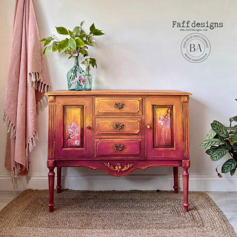 Bougainvillea Terra Clay Paint provides endless possibilities for your imagination and can be used on wood, canvas, tile, fabric and more!
Wet distressed credenza with Bougainvillea Terra Clay Paint on the bottom portion of the ombre with Daffodil Terra Clay Paint, and Marigold Terra Clay Paint as the top ombre colors. The Belles & Whistles Bud & Branches - Transfer can be seen on the inside door panels. Staged on top of a woven rug and a pink linen fringe blanket hanging behind piece.