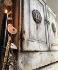 Terra Clay Paint Desert Tan Terra Clay Paint 
This is a very distressed wooden bench with two doors on the front.  There are appliques on both doors.  To the left is a lit, black candle, mesh, brown rope with oyster shells hanging off of it.  There is also a brown book and a branch on the floor.