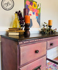 Elderberry Terra Clay Paint provides endless possibilities for your imagination and can be used on wood, canvas, tile, fabric and more! Pink and purple clay painted desk makeover with a dark stained top. Staged with a stack of 3 books with a yellow glass vase containing roses, a large candle stick with a purple candle, 2 candlesticks with orange candles and faux greenery. Large canvas painting on the wall over the desk of a woman with a colorful flower bouquet for hair.