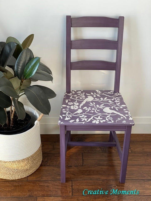 Elderberry Terra Clay Paint provides endless possibilities for your imagination and can be used on wood, canvas, tile, fabric and more! Purple clay painted chair with the Songbirds stencil applied in white paint. Large rubber plant in a white woven basket next to the chair.