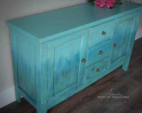 Lani's Lagoon Terra Clay Paint provides endless possibilities for your imagination and can be used on wood, canvas, tile, fabric and more!
Entertainment center cabinet painted in Lani's Lagoon Terra Clay Paint  as well as Blue Agave, Cerulean Blue Terra Clay Paint, Blue Moon and Malachite used in layers and a gradient between the colors. Staged on a white wall and gray floors.