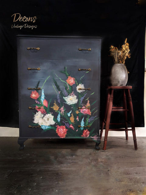 Wisteria Mist Terra Clay Paint provides endless possibilities for your imagination and can be used on wood, canvas, tile, fabric and more!  A black chalk painted dresser with a bouquet of flowers hand painted with Terra Clay Paint in assorted colors on the front.  The hardware is bronze.  A brown chalk painted wooden stool sits to the right of the dresser with a brown vase holding brown flowers.  Everything sits on a brown wooden floor, in front of a black and white wall.