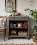 A green cabinet with two drawers and open shelves holds books, baskets, and decor. A plant, candles, and a framed leaf print sit on top. A wooden chair with a white cushion is on the left. A plant in a pot is on the right, on a textured rug.